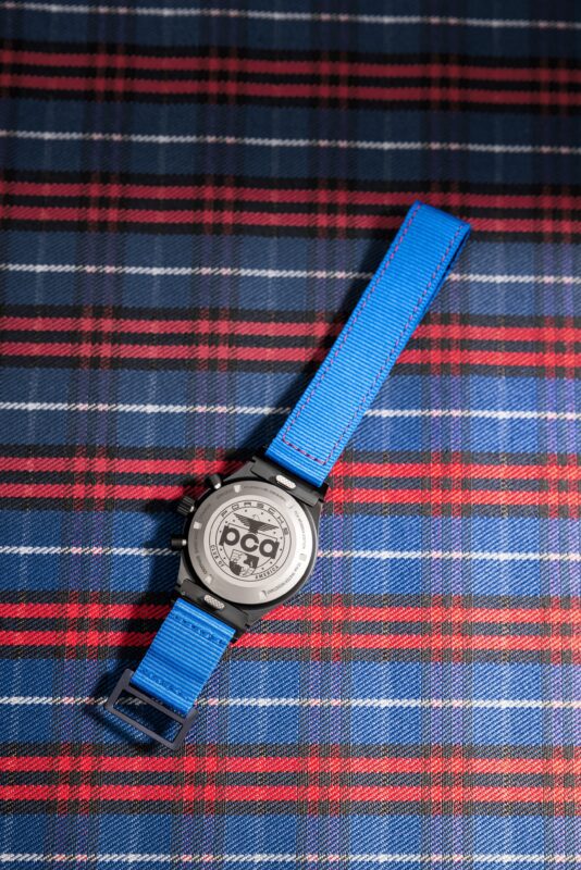 A Chronograph 1 by Porsche Design, featuring a blue nylon strap and engraved metal back, is placed face-down on red, blue, and white plaid fabric.