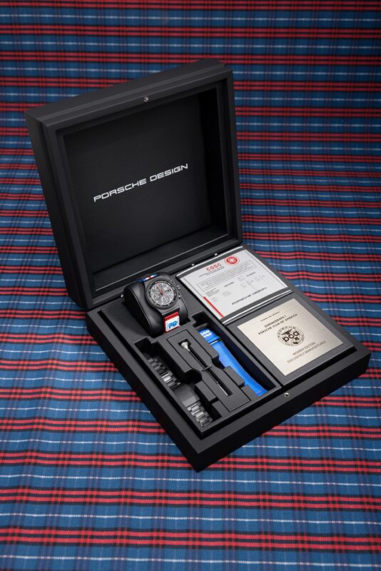 A Porsche Design Chronograph 1 watch set is displayed in an open black box with warranty cards, extra straps, and accessories, placed on a red and blue plaid surface—a perfect collector’s item for any PCA enthusiast.