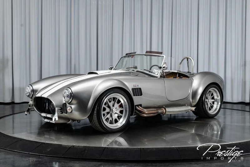 Silver Shelby Cobra replica with white racing stripes displayed on a rotating platform in a showroom with grey curtains—a standout among custom cars for sale.