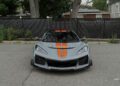 A gray 2025 Chevrolet Corvette ZR1 with orange racing stripes is parked on a paved street in front of a black fence and trees, showcasing its bold style and 064HP power. This impressive sports car could soon be yours—for sale now!.
