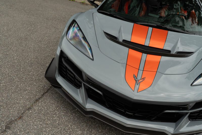A close-up of the front end of a gray 2025 Chevrolet Corvette ZR1 with orange racing stripes, parked on an asphalt surface.