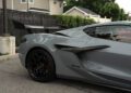 A close-up side view of a 2025 Chevrolet Corvette ZR1 with a large rear wing, black wheels, and visible air vents, parked on a street near houses and a fence—this sleek sports car is now for sale.