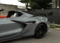 Close-up view of the rear side of a 2025 Chevrolet Corvette ZR1 with black wheels, a large rear spoiler, and orange interior, parked next to a black fence—this stunning sports car is now for sale.