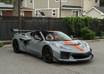 A gray 2025 Chevrolet Corvette ZR1 with black wheels and orange racing stripes is parked on a street in front of a black fence and houses, looking like it’s ready for sale to an eager new owner.