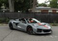 A gray 2025 Chevrolet Corvette ZR1 with black and orange accents is parked on a street near a black fence and trees.