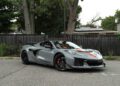A gray 2025 Chevrolet Corvette ZR1 with orange accents and 1064HP is parked on a street in front of a black fence and trees, ready for sale.