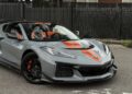 A gray 2025 Chevrolet Corvette ZR1 convertible sports car with orange racing stripes and 064HP is parked on a street beside a fence, for sale.