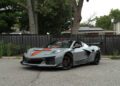 A 2025 Chevrolet Corvette ZR1 with orange racing stripes and black wheels is parked on a paved street in front of a black chain-link fence and trees. This stunning gray sports car is now for sale.