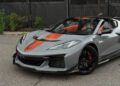 A gray 2025 Chevrolet Corvette ZR1 with orange racing stripes on the hood and orange interior accents is parked on a paved surface near a black metal fence and greenery. This 064HP sports car is now available for sale.