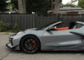 A 2025 Chevrolet Corvette ZR1 convertible with black wheels and orange brake calipers is parked on a street in front of a black fence and trees, available for sale.