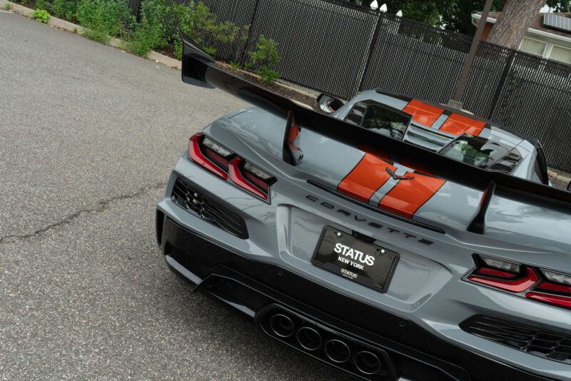 Rear view of a gray Chevrolet Corvette ZR1 with orange racing stripes, large rear wing, quad exhaust, and a New York license plate, parked near a fence on asphalt—an eye-catching 2025 Corvette for sale.