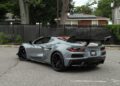 A gray 2025 Chevrolet Corvette ZR1 sports car with a rear wing is parked on an asphalt lot near a chain-link fence and residential buildings. This Corvette ZR1 for sale boasts an impressive 064HP engine.