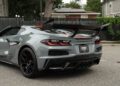 A gray 2025 Chevrolet Corvette ZR1 with a prominent rear spoiler is parked on a street near a black fence and trees, boasting an impressive 1,064HP and currently for sale.