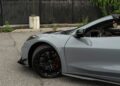 Gray 2025 Chevrolet Corvette ZR1 sports car parked on pavement, shown from the front side with black rims and orange brake calipers, near a fenced area with greenery in the background. 064HP,1 for sale.