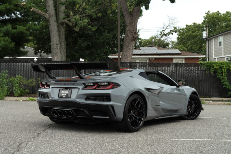 A gray 2025 Chevrolet Corvette ZR1 sports car is parked in an outdoor lot next to trees, a fence, and residential buildings, and is now for sale.