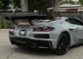 A gray 2025 Chevrolet Corvette ZR1 with a large rear spoiler is parked on a paved surface near a fence and trees.