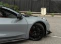 A close-up of the front left side of a 2025 Chevrolet Corvette ZR1 with black wheels, parked in a lot near a fence and some plants—showcasing its striking design and 1064HP performance, possibly for sale.
