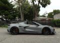 A gray 2025 Chevrolet Corvette ZR1 convertible sports car with black wheels is parked on a street next to a black fence and trees; this 1,064HP beauty could be yours, as it's now for sale.