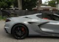A 2025 Chevrolet Corvette ZR1 with 064HP, black alloy wheels, orange brake calipers, and a raised rear wing is parked on a paved surface near a fence and trees—this stunning sports car is now for sale.