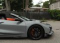 A gray 2025 Chevrolet Corvette ZR1 convertible with black wheels and orange accents is parked on a street near a black fence and residential houses.