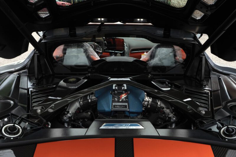 Close-up view of a 2025 Chevrolet Corvette ZR1 engine bay, showcasing a clean, high-performance 1,064HP engine with carbon fiber components and orange accents, with the car’s interior visible in the background.