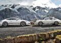 Two white 2016 Porsche 911 R sports cars parked side by side on a mountain road, framed by snow-covered peaks and rugged, rocky terrain in the background.