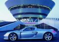 A silver Porsche Carrera GT is parked in front of a modern, circular building with "Porsche" signage, likely at a Porsche facility—a perfect tribute to the legendary Carrera GT and its enduring appeal.
