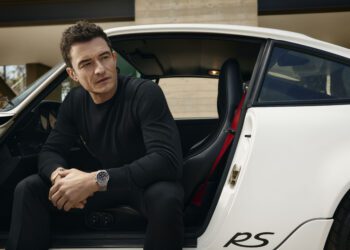A man in a black outfit, reminiscent of Orlando Bloom’s sleek style, sits in the open doorway of a white car with "RS" lettering, looking to the side with a neutral expression.