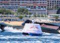 A person steers a sleek black Porsche Macan EV-Powered Frauscher Fantom 850 near a floating buoy with "Yacht Club de Monaco" written on it, with a city and beachfront in the background as it sets a speed record in Monaco.