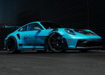 A PTS Ipanema Blue 911 GT3 RS from Porsche Exchange is parked on a blacktop in front of a dark, ribbed metal wall.