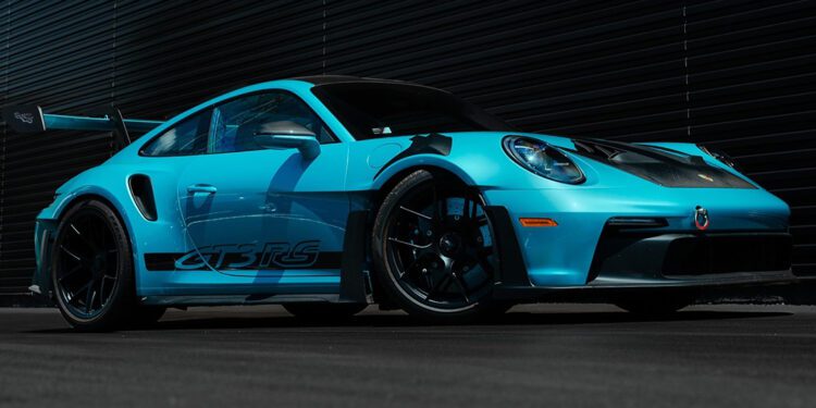 A PTS Ipanema Blue 911 GT3 RS from Porsche Exchange is parked on a blacktop in front of a dark, ribbed metal wall.
