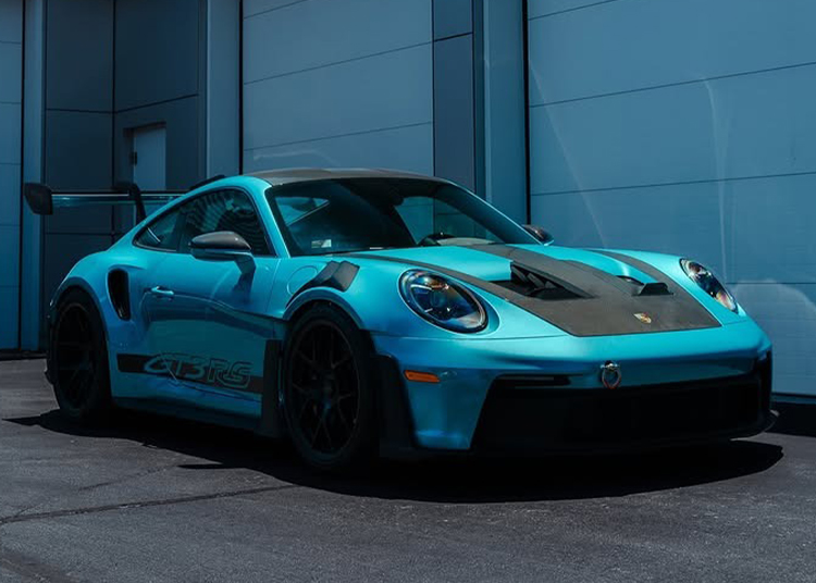 A Porsche Exchange 911 GT3 RS in PTS Ipanema Blue with a rear spoiler is parked in front of a modern building clad in light blue panels.