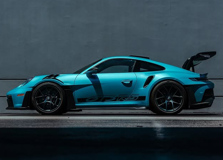 An Ipanema Blue 911 GT3 RS with a rear spoiler is parked on a street against a plain gray wall, viewed from the side. Discover this Porsche at Porsche Exchange.