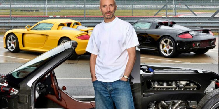 A man in a white t-shirt and jeans stands in front of three high-performance Porsche sports cars, including a Carrera GT, on a racetrack with a chain-link fence—capturing the essence of speed and style like a tribute capsule collection.