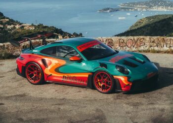 A green and orange Porsche 911 GT3 RS with a rear wing is parked on a scenic overlook, with a coastal town and sea in the background—looking like it could headline an RM Sotheby's Auction or feature in Dua Lipa's next music video.