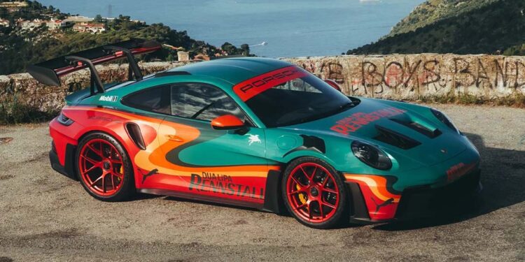 A green and orange Porsche 911 GT3 RS with a rear wing is parked on a scenic overlook, with a coastal town and sea in the background—looking like it could headline an RM Sotheby's Auction or feature in Dua Lipa's next music video.