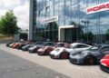 A row of race-ready Porsche sports cars, including the GT3 RS, is parked in front of a glass building with "Manthey" and "Porsche" signage on the facade.