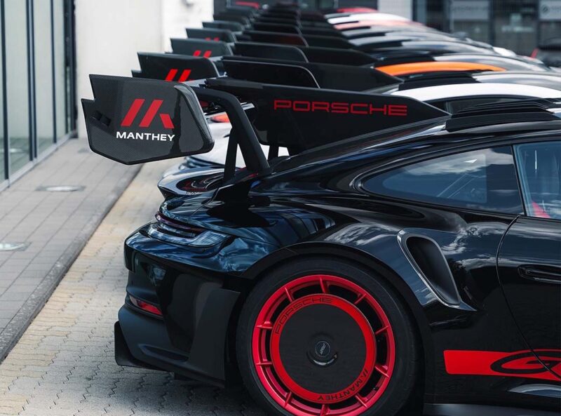 A row of black Porsche sports cars with red accents is parked, featuring large rear wings with "Manthey" and "Porsche" branding, highlighting their race-ready GT3 RS MR specs from Manthey Racing.
