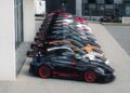 A row of race-ready Porsche GT3 RS sports cars, mostly black, white, and silver with large rear spoilers, parked diagonally beside a modern building.