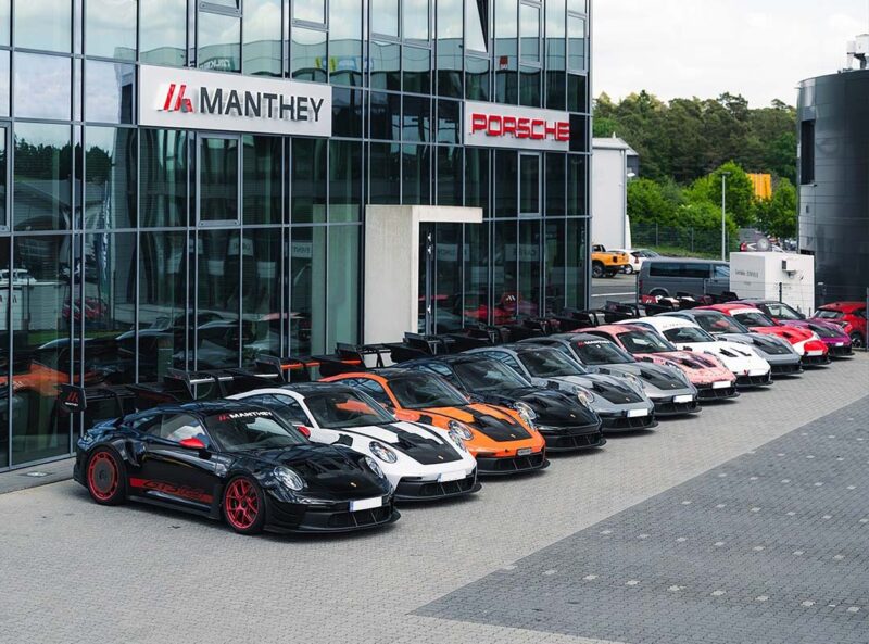 A row of race-ready Porsche sports cars, including GT3 RS models in various colors, is parked outside a building with Manthey Racing and Porsche signage.
