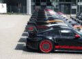 A row of race-ready black Porsche 911 GT3 RS MR cars with red accents and large rear wings are parked on a paved lot.
