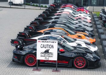 A row of parked Porsche sports cars with large rear wings, including a GT3 RS MR, sits behind a sign reading "Manthey Caution Shark Parking Zone," showcasing Manthey Racing’s race-ready machines.
