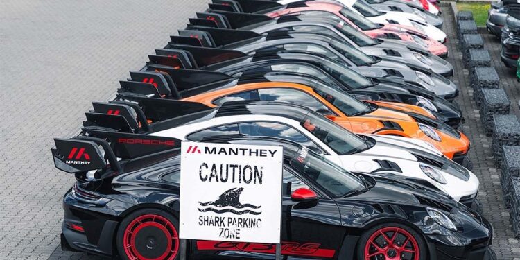 A row of parked Porsche sports cars with large rear wings, including a GT3 RS MR, sits behind a sign reading "Manthey Caution Shark Parking Zone," showcasing Manthey Racing’s race-ready machines.