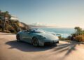 A silver Porsche 911 convertible sports car is parked on a sunlit coastal road with ocean waves, cliffs, and a modern house in the background.