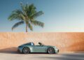 A green Porsche 911 convertible sports car is parked in front of an orange textured wall with a tall palm tree in the background under a clear sky.