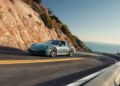 A silver Porsche 911 sports car drives along a winding coastal road bordered by rocky cliffs, with the ocean visible in the background under a clear sky.