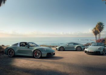 Three silver Porsche 911 variants are parked on a coastal road lined with palm trees, overlooking the ocean beneath a clear sky.