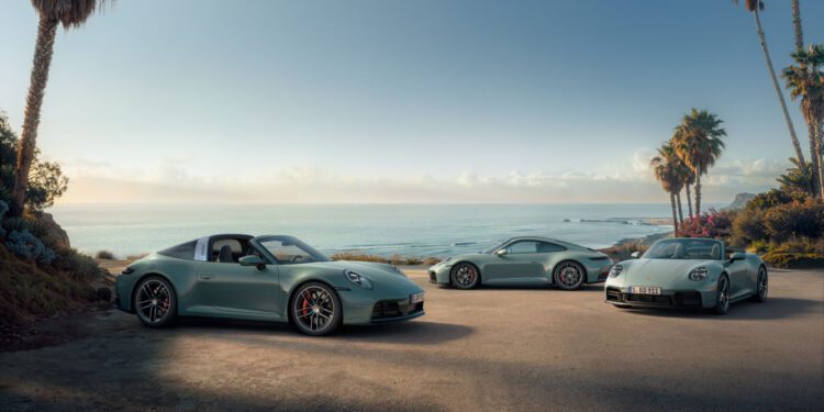 Three silver Porsche 911 variants are parked on a coastal road lined with palm trees, overlooking the ocean beneath a clear sky.