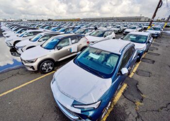 Rows of new cars are parked in a large outdoor lot on an overcast day, with puddles reflecting the vehicles—poised for export as the Trump von der Leyen landmark deal aims to boost U.S. auto exports to Europe by slashing tariffs.
