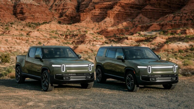 Two green Rivian Gen 2 electric vehicles, the R1T Quad-Motor pickup and an SUV, are parked on a dirt road with stunning red rock formations in the background.